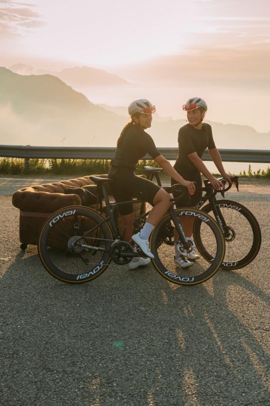 Dos ciclistas en una carretera con un sofá al fondo durante el atardecer