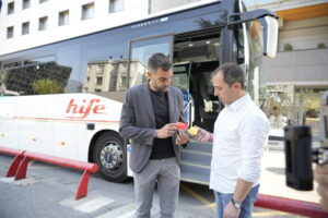 Dos hombres revisan tarjetas de autobús frente a un vehículo de transporte público.