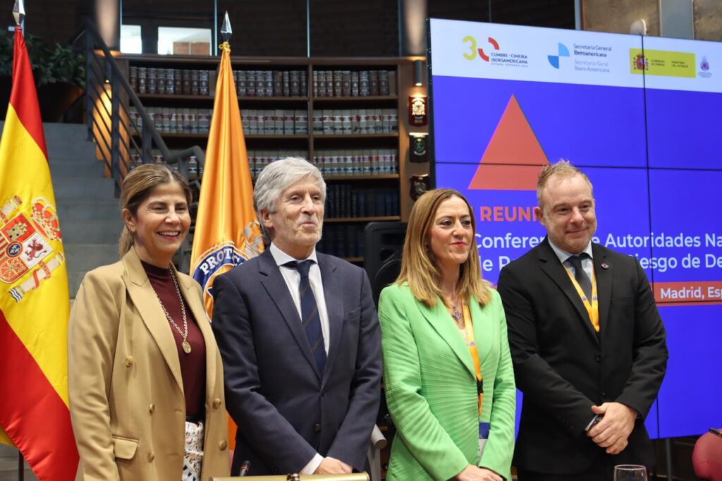 Cuatro asistentes posan ante banderas y una pantalla de una reunión sobre protección civil.