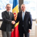 Dos hombres estrechan la mano frente a una bandera de Andorra y una escultura.