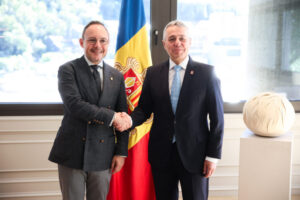 Dos hombres estrechan la mano frente a una bandera de Andorra y una escultura.
