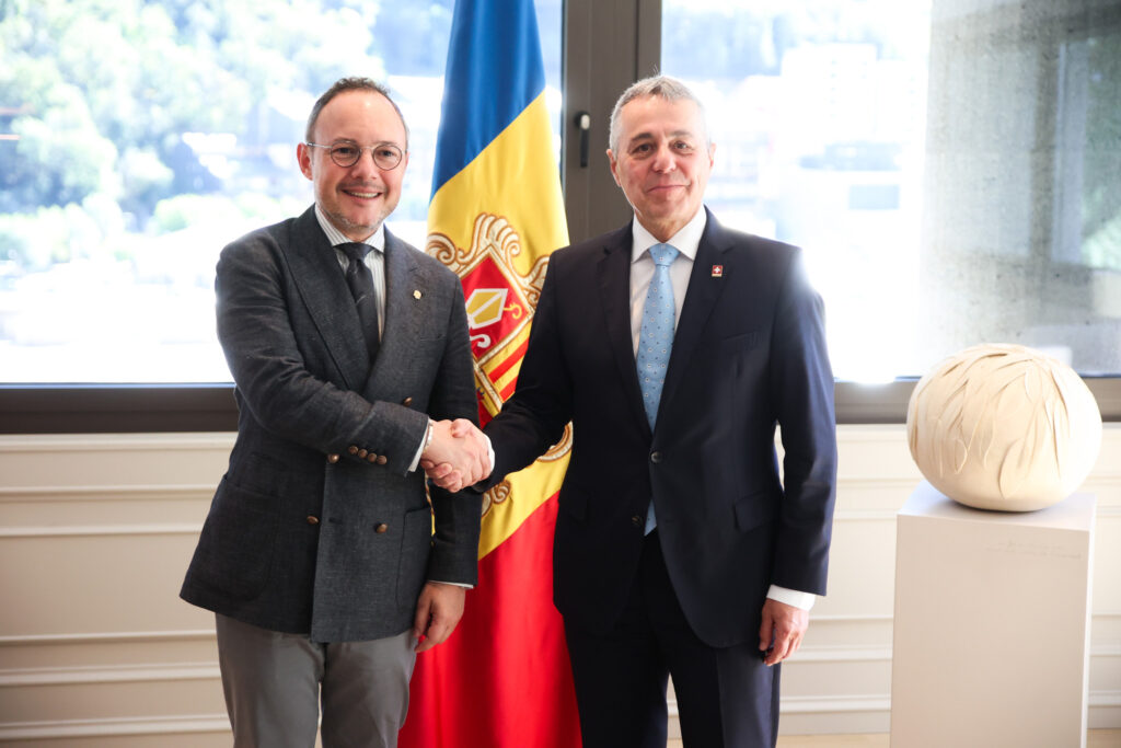 Dos hombres estrechan la mano frente a una bandera de Andorra y una escultura.
