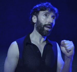 Fotografía de un hombre con chaleco negro alzando el puño bajo iluminación azul en escena teatral.