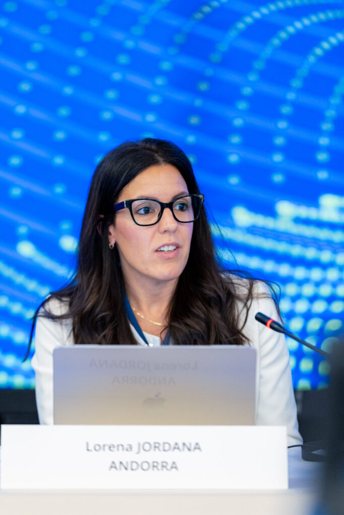 Lorena Jordana sentada ante un portátil y un micrófono, con un cartel de Andorra
