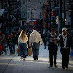 Varias personas caminan por una calle comercial iluminada y algunas llevan bolsas de compras.