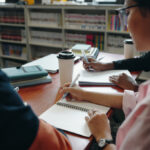 Varias personas estudian y escriben en cuadernos alrededor de una mesa con libros.