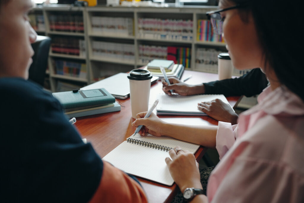 Varias personas estudian y escriben en cuadernos alrededor de una mesa con libros.