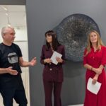Tres personas están de pie frente a una obra circular en una galería.