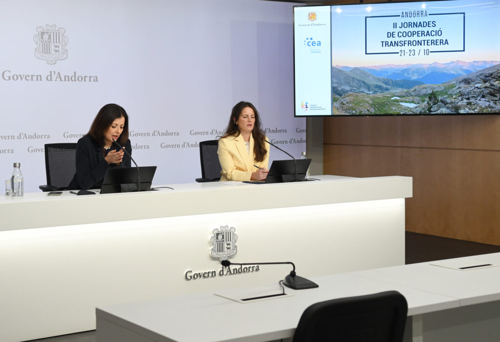 Dos mujeres sentadas ante una mesa con portátiles en una sala institucional.